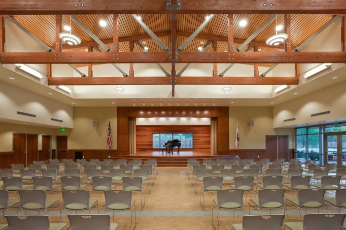 Inside view of large event center with stage.  Baby grand piano is on the stage and chairs are set in rows with a center isle theater style.
